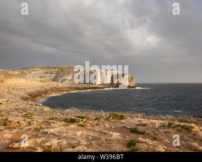 Bella round bay, piante verdi e Fungus Rock Cliffs al tramonto a Gozo. Arancione della luce al tramonto con il grigio nuvole temporalesche. Foto Stock