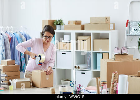 Bruna giovane donna scatola di tenuta con ordine online di client con cellotape mentre lavoro in ufficio Foto Stock