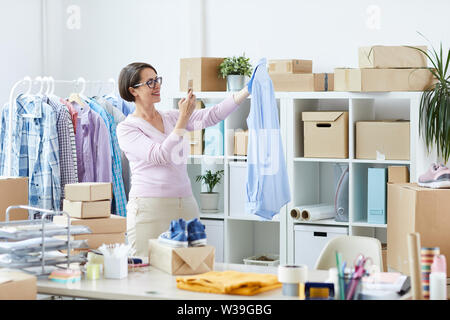 Lavoratore di ufficio con lo smartphone tenendo immagine del nuovo elegante maglietta blu su appendiabiti per uno dei client Foto Stock