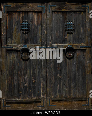 Primo piano di una doppia porta in legno intemprata con maniglie ad anello in metallo e piccole finestre barrate in uno stile rustico e antico Foto Stock