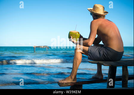 Tourist indossando una paglia cappello per il sole seduto con una noce di cocco verde su un rustico in legno Tavolo picnic in corrispondenza di un'isola tropicale beach Foto Stock