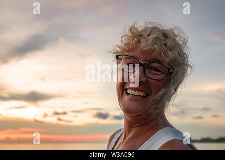 Un anziano conciate donna felice sorride al tramonto in riva al mare sulla spiaggia. aria aperta, sera, close-up. Foto Stock