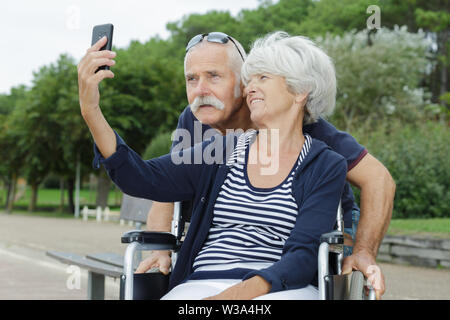 Coppia senior in stazionamento tenendo selfie donna in sedia a rotelle Foto Stock