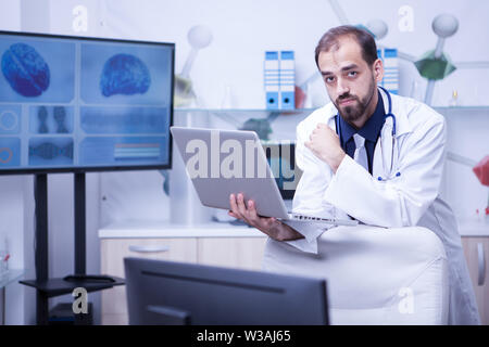 Giovane medico barbuto guardando la telecamera e in possesso di un computer portatile. Medico utilizzando la tecnologia. Foto Stock