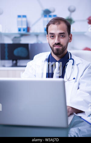 Ritratto di giovane medico che lavorano su laptop e sorridente tot la fotocamera. Medico con lo stetoscopio intorno al suo collo. Foto Stock