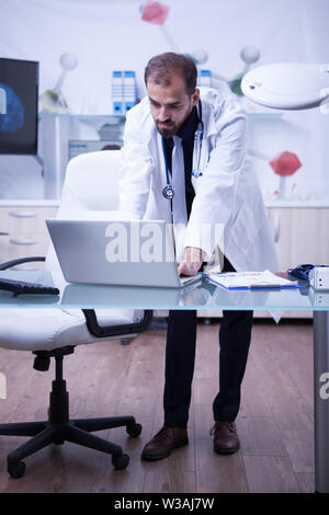 Full lenght ritratto di bello medico al lavoro sul suo computer portatile presso l ospedale mobile. Dottore in un laboratorio. Foto Stock