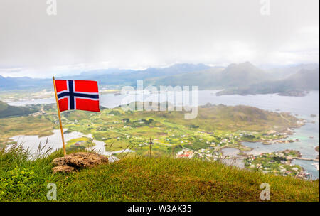 Norwegian bandiera nazionale nel vento sulla sommità del picco Nonstinden con fjord in background, Ballstad, Vestvagoy comune, Nordland county, né Foto Stock