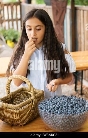 Ragazza giovane snacking da una ciotola di appena raccolto sano di mirtilli maturi su un tavolo all'aperto in estate Foto Stock