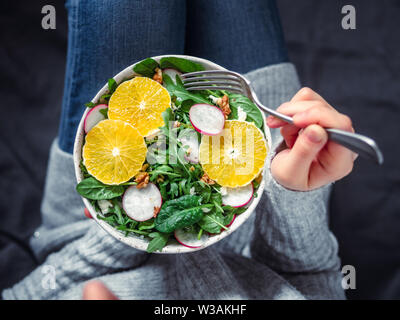 Donna in jeans a letto, azienda vegan insalatiera con arance, spinaci,la rucola,ravanello,dado.Top view.Vegan colazione,cibo vegetariano,la dieta concetto.ragazza in jeans forcella di supporto con le ginocchia e le mani visibili Foto Stock