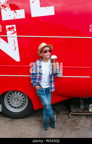 Little Boy in occhiali da sole e cappello è bere latte cocktail rosso su sfondo sicy Foto Stock