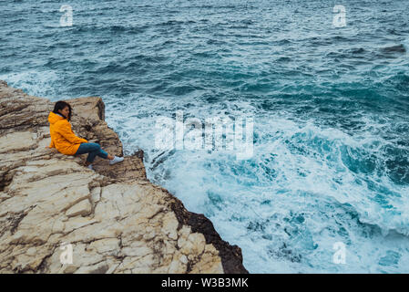 La donna posa sul bordo della scogliera alla ricerca sulle grandi onde del mare Foto Stock