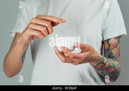 Donna tatuata mani tenendo un vasetto di crema. Dito con una goccia di crema bianca Foto Stock