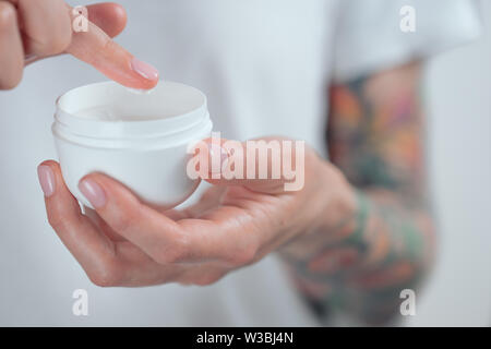 Donna tatuata mani tenendo un vasetto di crema. Dito con una goccia di crema bianca Foto Stock