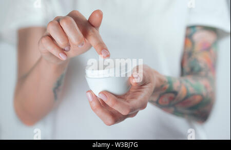 Donna tatuata mani tenendo un vasetto di crema. Dito con una goccia di crema bianca Foto Stock