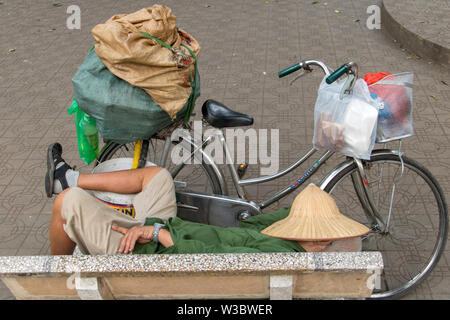 HANOI, Vietnam, Apr 21 2019, un uomo si rilassa su un banco con un cappello sulla sua testa e una bicicletta con molti sacchi. Foto Stock