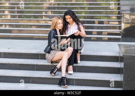 Due belle donne gli studenti di diverse nazionalità sono la lettura di un libro seduti sui passi dell'università Foto Stock