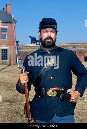 Un uomo barbuto che indossa la divisa militare di U.S. La guerra civile europea soldato pone con armi che egli porta come un interprete che accoglie i visitatori a Fort Clinch parco dello stato in Fernandina Beach sulla costa orientale della Florida, Stati Uniti d'America. La sua mano destra trattiene un Springfield rifled moschetto con il suo allegato a baionetta e una pistola è infilata nella cintura di cuoio intorno ai lombi che porta anche le munizioni. Questo storico fort all'entrata di Cumberland Sound e la Basilica di Santa Maria del fiume è stata inizialmente occupata dall'esercito confederato fino a quando passò sotto il controllo delle truppe dell'Unione nel 1862. Foto Stock