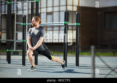 Montare il bel giovane uomo barbuto con coda di cavallo accovacciato e alzando le mani direttamente praticando esercizio di stiramento all'aperto Foto Stock