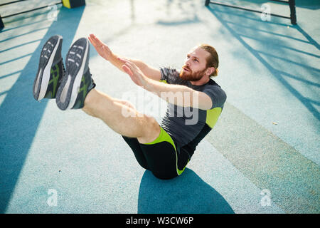Concentrato giovane uomo barbuto in sportswear seduta sul terreno esterno e facendo sgranocchia con gambe sollevate Foto Stock