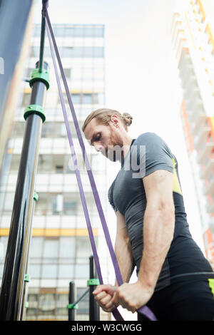 Forte concentrato giovane uomo barbuto con coda di cavallo in piedi in attrezzature sportive e tirando verso il basso tendere la cinghia Foto Stock