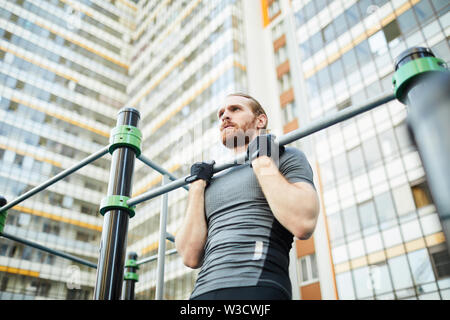 Grave determinata giovane uomo barbuto onu guanti senza dita esecuzione di pull-up su sport bar contro l edificio di appartamenti Foto Stock