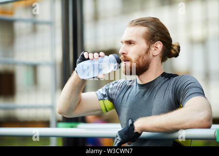 Contemplativo bel giovane uomo barbuto con coda di cavallo appoggiata sulla barra e acqua potabile per recuperare la forza Foto Stock