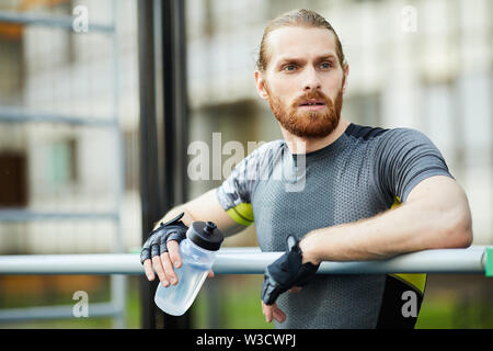 Pensieroso giovane sportivo barbuto in sport sudate shirt appoggiata sulla barra e acqua potabile mentre facendo rompere Foto Stock