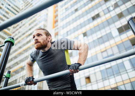 Apposite giovane uomo barbuto in sport tshirt guardando dritto e facendo pull-ups sulla barra contro edificio di appartamenti Foto Stock