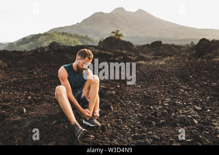 Giovane uomo seduto e legatura di scarpe da corsa prima di trail running all'esterno. Paesaggio vulcanico e vista sulle montagne sullo sfondo. Foto Stock