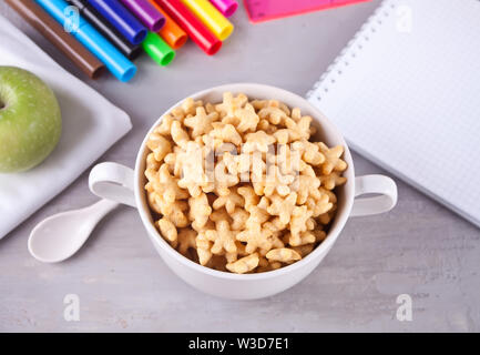 Una ciotola di stelle secca Cereali, mela verde, colorati pennarelli e notebook sul tavolo grigio per la salute la prima colazione per gli adulti e i bambini. Vista dall'alto. Scho Foto Stock