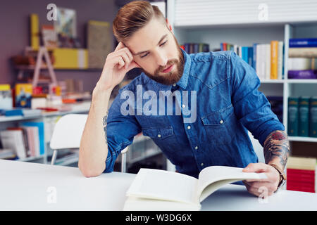 Sottolineato maschile giovane studente la lettura di un libro di testo mentre è seduto nella libreria Foto Stock