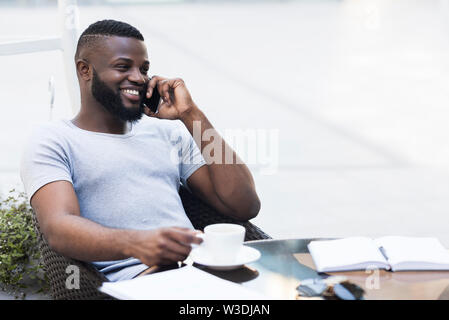 Giovane imprenditore africano rilassante in un cafe e sorridente Foto Stock