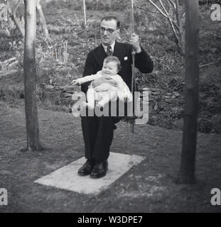 anni '50, storico, un uomo che indossa un vestito e cravatta seduta su un'oscillazione di corda in un giardino che tiene un bambino infante, non cercando confortevole. Foto Stock