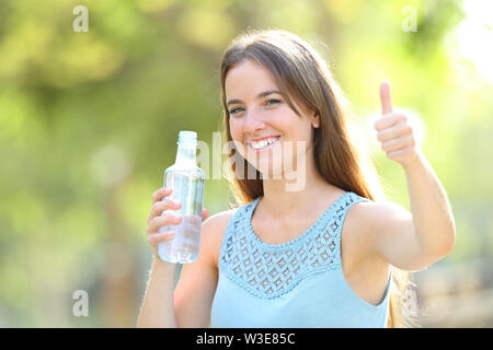 Donna felice tiene una bottiglia di acqua con il pollice in su in un parco con uno sfondo verde Foto Stock