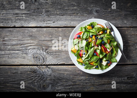 Insalata estiva con fresche foglie di bietole, pesche, mirtilli, pezzi di formaggio con colore verde-bluastro vene servita su una piastra bianca su una tavola in legno rustico, h Foto Stock