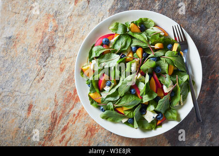 Deliziosa insalata sana con fresche foglie di bietole, pesche, mirtilli, pezzi di formaggio con colore verde-bluastro vene servita su una piastra bianca, vista orizzontale Foto Stock