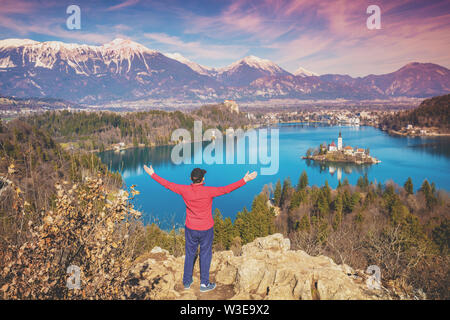 Uomo con le mani in aria in piedi su una scogliera oltre il lago di Bled in primavera. La Slovenia, Europa Foto Stock