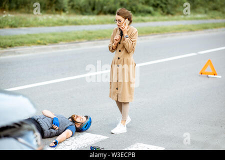 Incidente stradale con feriti ciclista e preoccupati driver donna chiamando con telefono sul passaggio pedonale vicino alla rotta per biciclette e auto Foto Stock