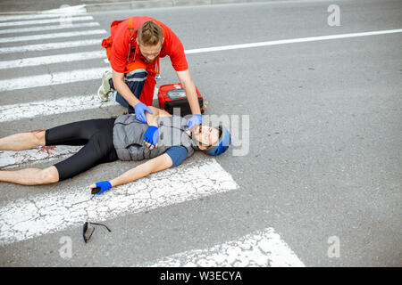 Medic l'applicazione di primo soccorso ai feriti ciclista giacente sul passaggio pedonale dopo l'incidente stradale Foto Stock