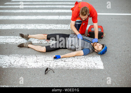 Medic l'applicazione di primo soccorso ai feriti ciclista giacente sul passaggio pedonale dopo l'incidente stradale Foto Stock