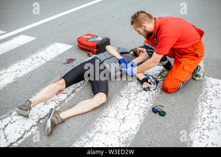 Medic l'applicazione di primo soccorso ai feriti ciclista giacente sul passaggio pedonale dopo l'incidente stradale Foto Stock