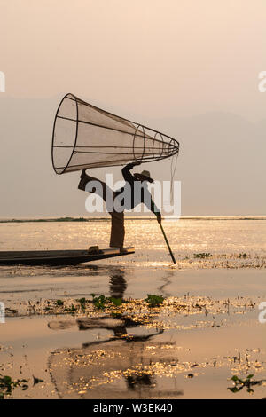 Inle, Myanmar - Marzo 2019: tradizionale birmana gamba pescatore di canottaggio a Lago Inle durante il sunrise. Silhouette di retroilluminazione riflesso nell'acqua Foto Stock