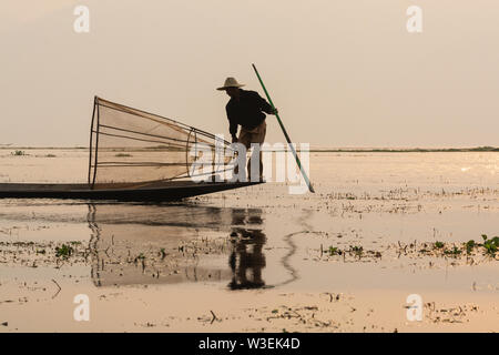 Inle, Myanmar - Marzo 2019: tradizionale birmana gamba pescatore di canottaggio a Lago Inle durante il sunrise. Silhouette di retroilluminazione riflesso nell'acqua Foto Stock