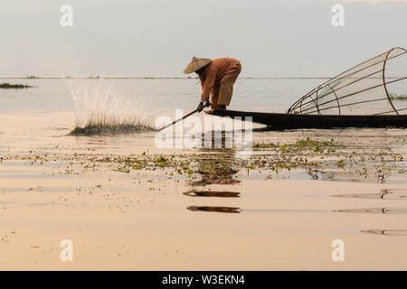 Inle, Myanmar - Marzo 2019: tradizionale birmana gamba pescatore di canottaggio a Lago Inle durante il sunrise smashing acqua con paddle Foto Stock