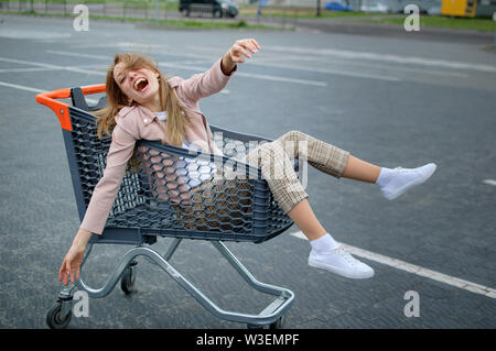 Crazy giovane ragazza in abiti casual e bianco sneaker scarpe nel parcheggio del supermercato divertirsi seduti nel carrello. Foto Stock