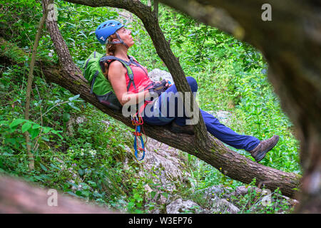 Donna alpinista con un casco blu in appoggio sul ramo di un albero, in Tureni-Copaceni gorge, Romania. La sua scalata marcia (sosta tubo, prusik loop carabina, Foto Stock