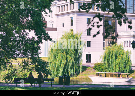 Due ragazze a sedersi su una panchina nel parco di Riga dalla citta di canale con una barca in estate Foto Stock