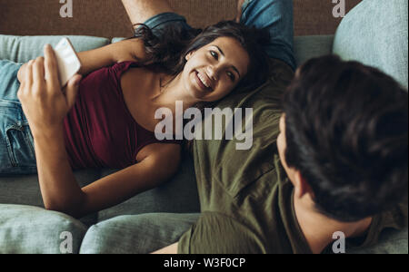 Giovane donna con smart phone giacente su fidanzati lap e sorridente. Giovane rilassante sul divano di casa. Foto Stock
