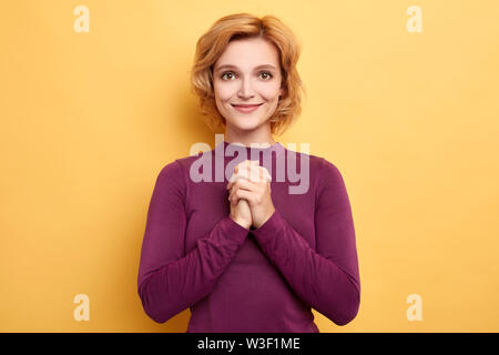 Sognante giovane donna affascinante mani tiene premuti insieme sotto il mento, guarda con felice espressione , ha sorriso toothy, indossa eleganti maglione viola , è Foto Stock