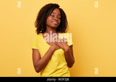 Romantico brunette ragazza con gli occhi chiusi, mantiene le palme sul cuore, sente gratitudine,vestiti in stile casual giallo t shirt, isolato su giallo backgrou Foto Stock